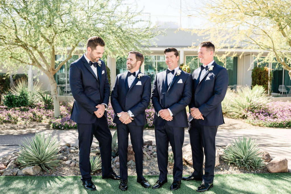 Groom and groomsmen standing in a line smiling, wearing black suits.
