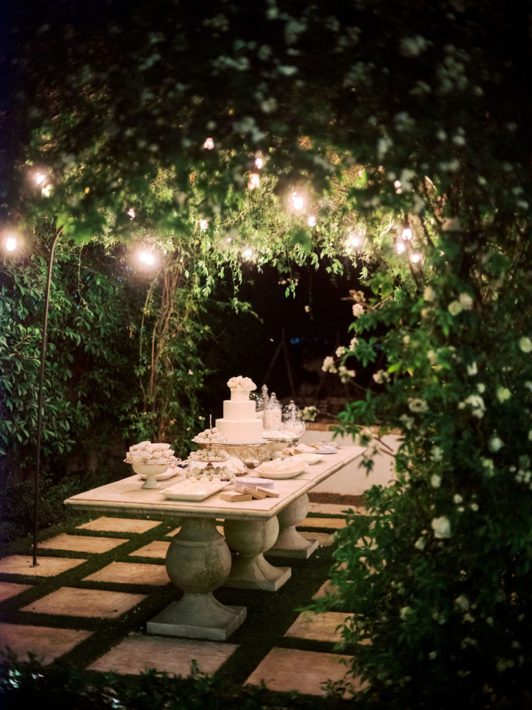 Romantic dessert table with cake and other items at wedding reception under outdoor greenery tunnel with white hanging lights above.