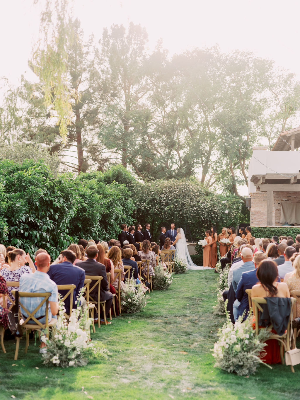Outdoor wedding ceremony on grass with wood chairs for guests and ground floral arrangements. Bride and groom at front.