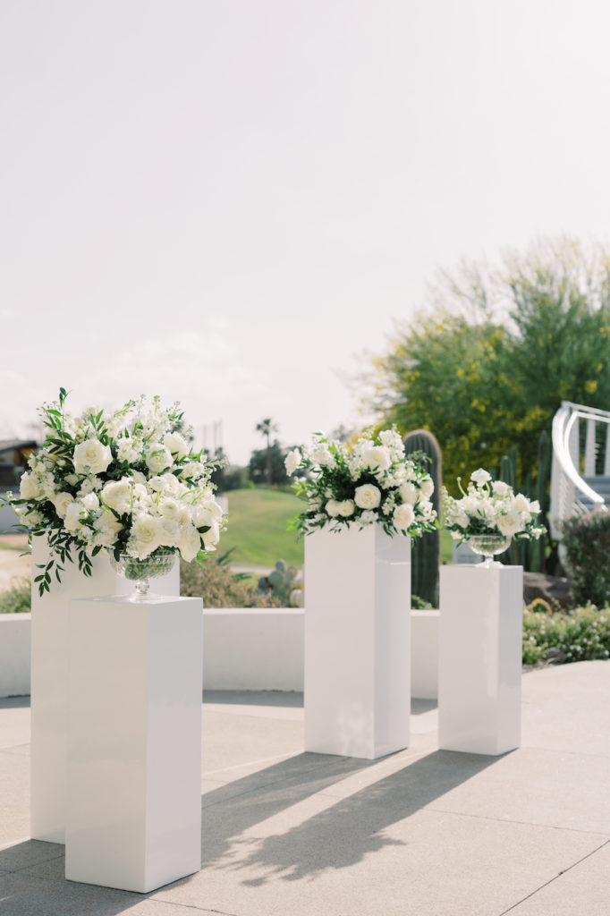 Wedding ceremony floral design of four floral arrangements of white flowers on white acrylic pedestals.