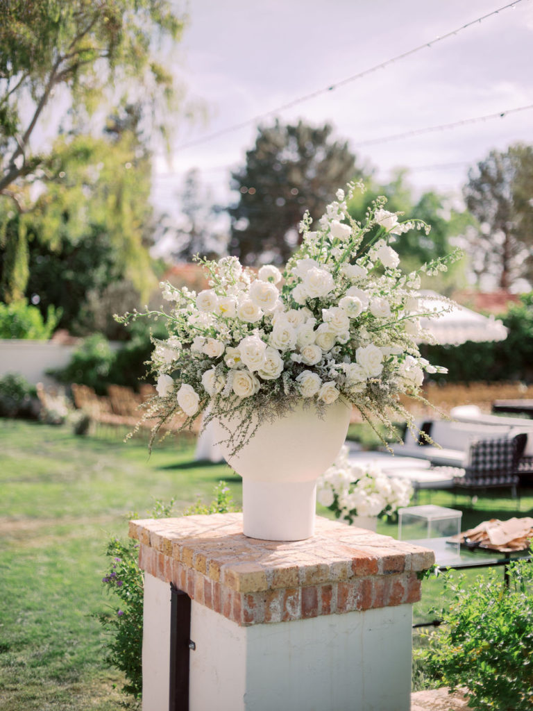 Large floral arrangement in large white vase filled with garden style white flowers and greenery set on brick pillar at wedding ceremony entrance.