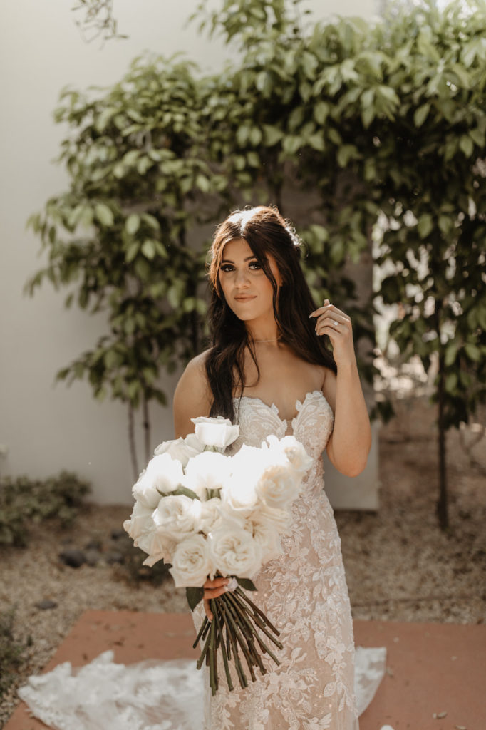 Bride holding long stem white roses bouquet, posing in greenery courtyard.