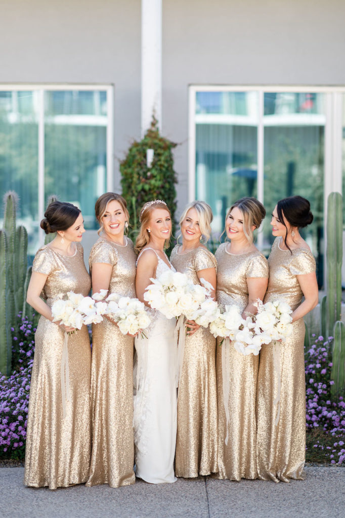 Bride standing with bridesmaids in gold dresses, all holding white floral bouquets