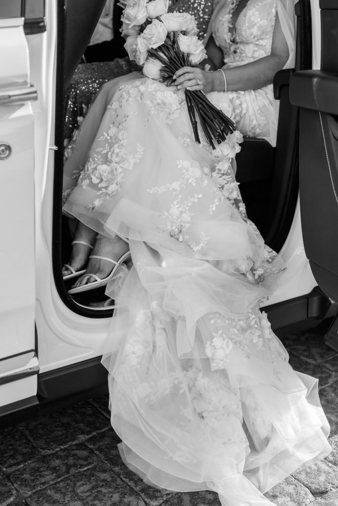 Bride holding long stem white roses sitting in vehicle with train trailing out.