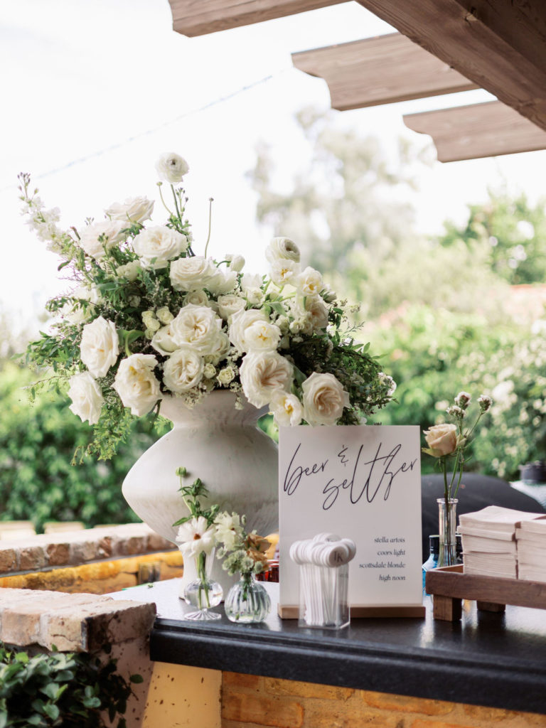 Large white floral and greenery arrangement in unique vase shape with bud vases set on wedding bar.