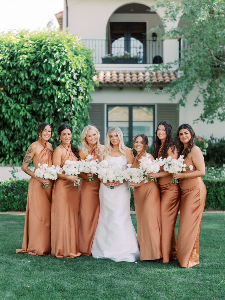 Bride standing in row with bridesmaids in copper dresses, all holding white floral bouquets.
