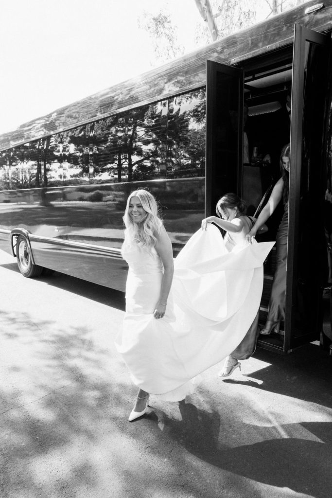 Bride exiting party bus with train lifted behind her by others.