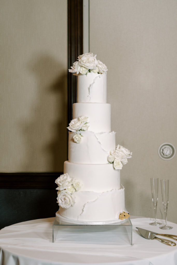 Five tier white wedding cake with white roses and small dog accent at bottom.