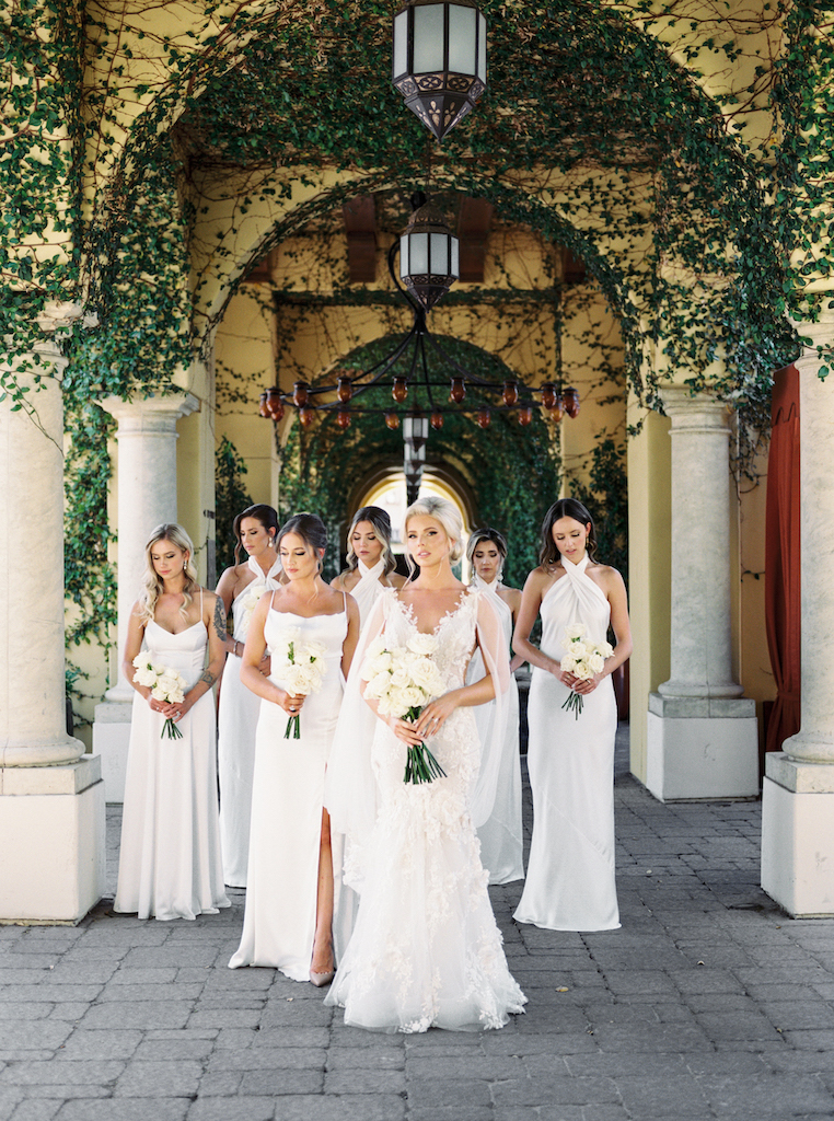 Bride standing with bridesmaids all in white under ivy covered outdoor walkway holding white rose bouquet.