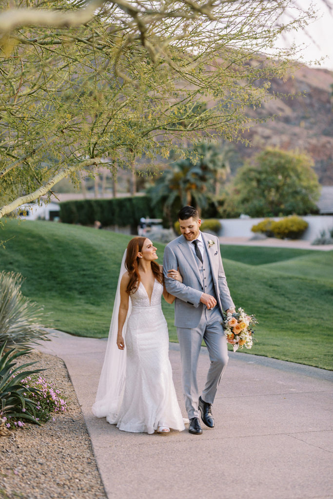 Bride walking with groom, arm in arm, at Mountain Shadows on cement sidewalk.
