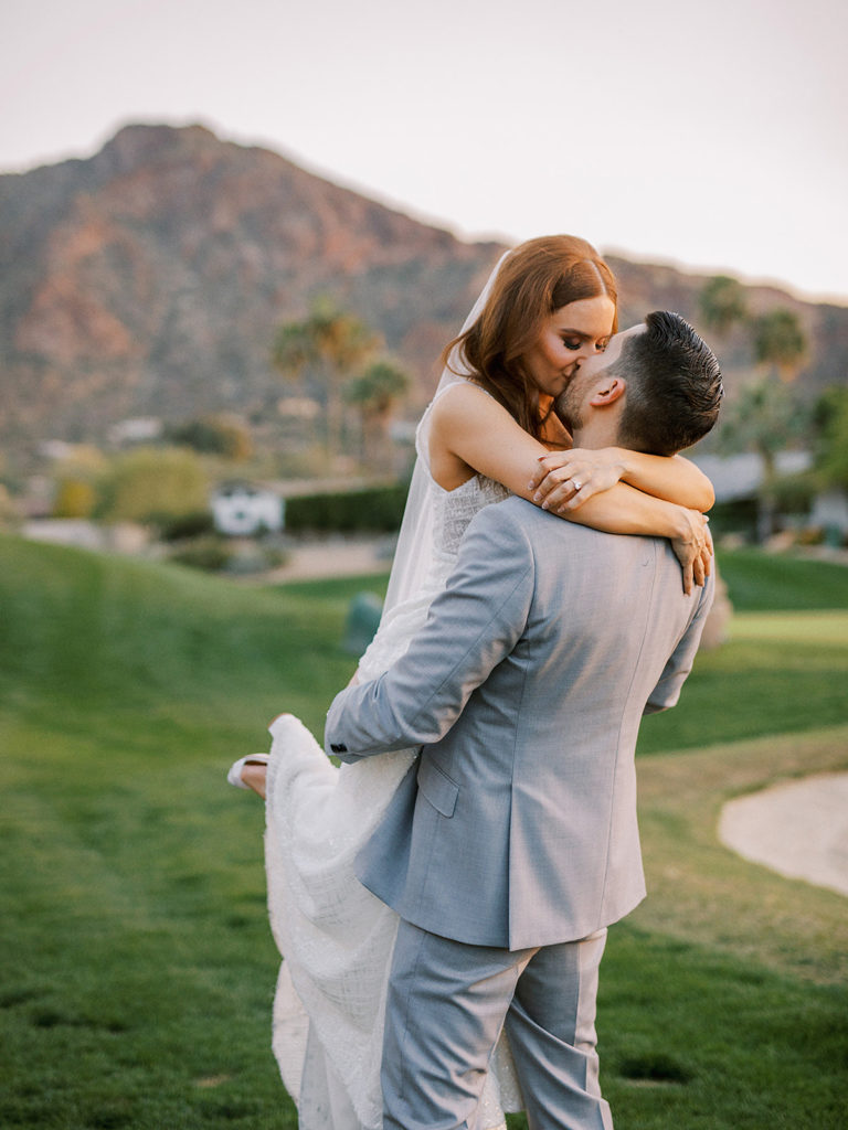 Groom holding up bride during kiss on golf course with desert mountain in background.