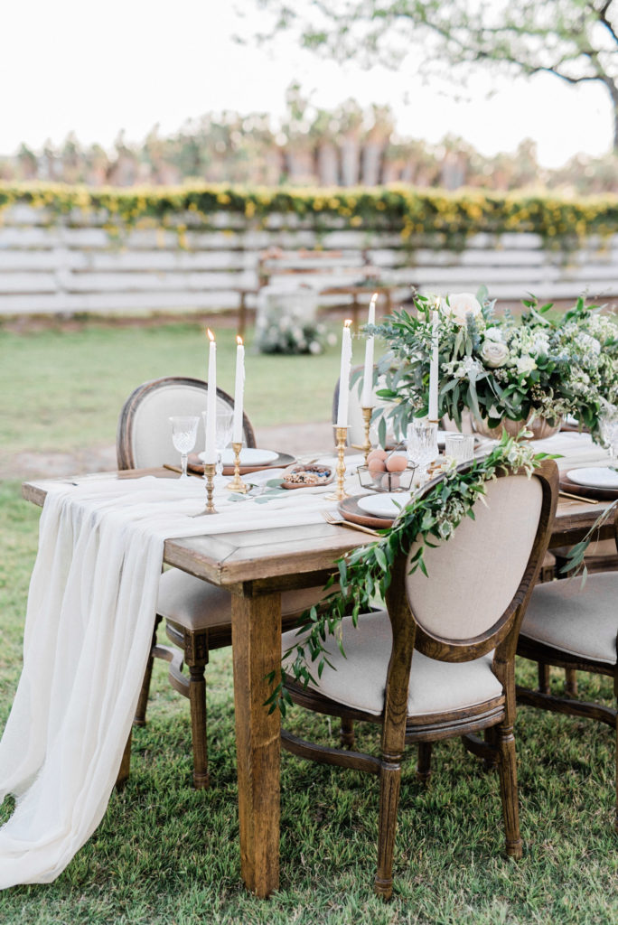 Set wooden table in grass field with white linens and white taper candles and floral.