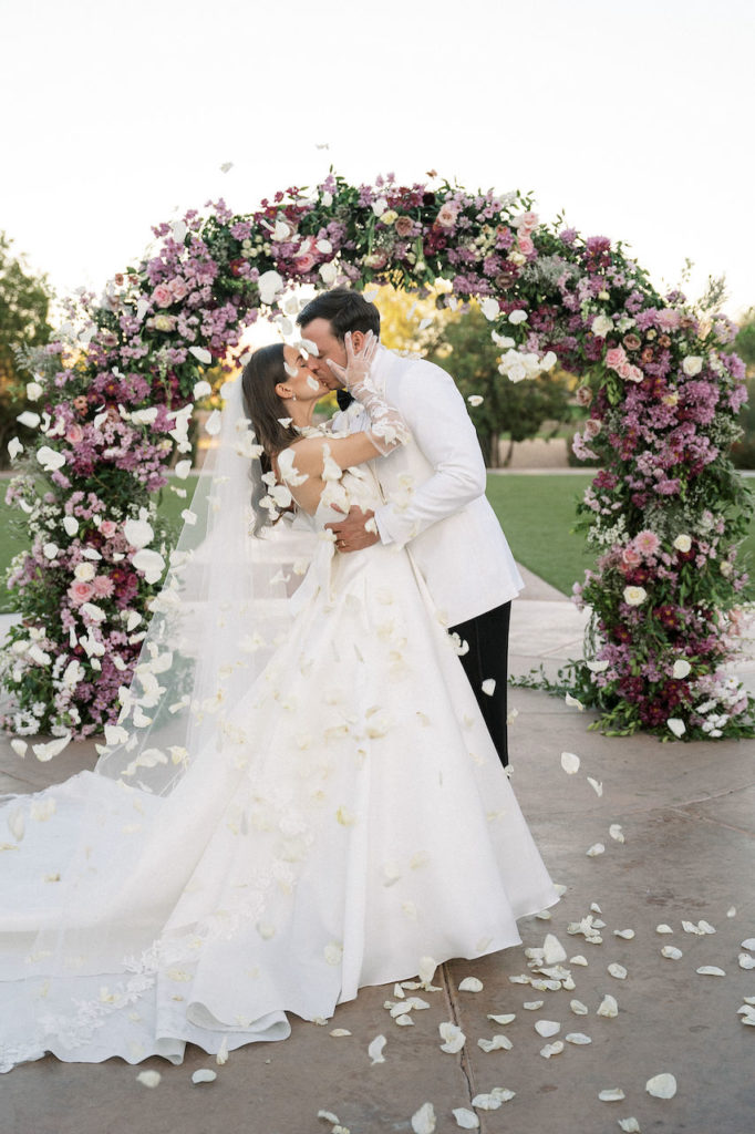 Bride and groom kissing during petal toss in front of wedding ceremony arch.