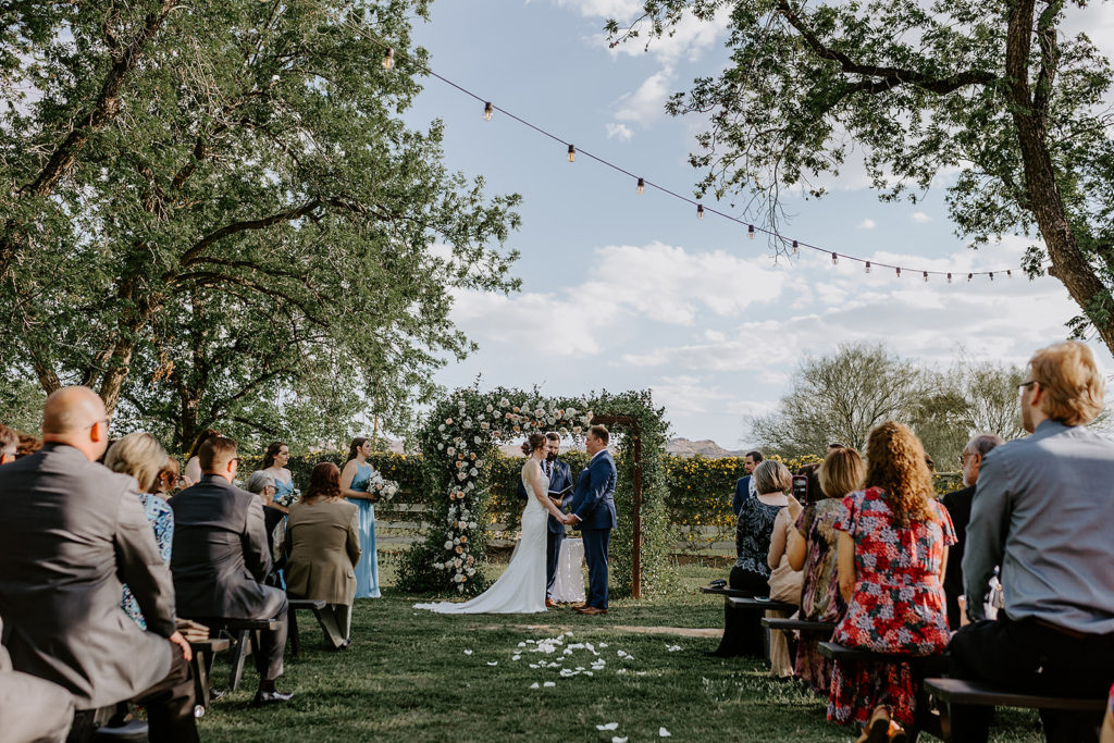 Bride and groom holding hands at outdoor wedding ceremony with officiant in front of arch of greens and white and pink floral.