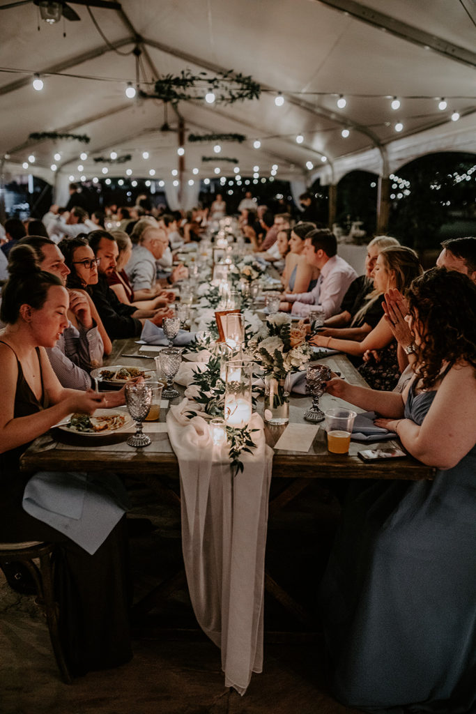 Wedding reception outdoor under tent of long rectangular tables with candles and greenery down the middle.