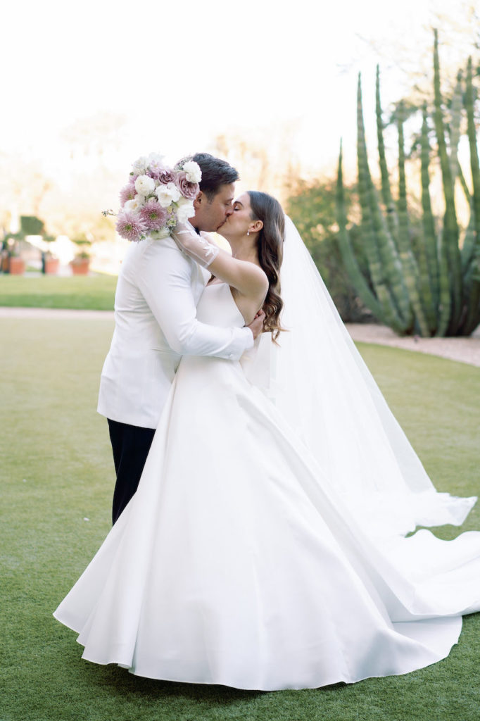 Bride and groom kissing in open grass area with cacti in background.