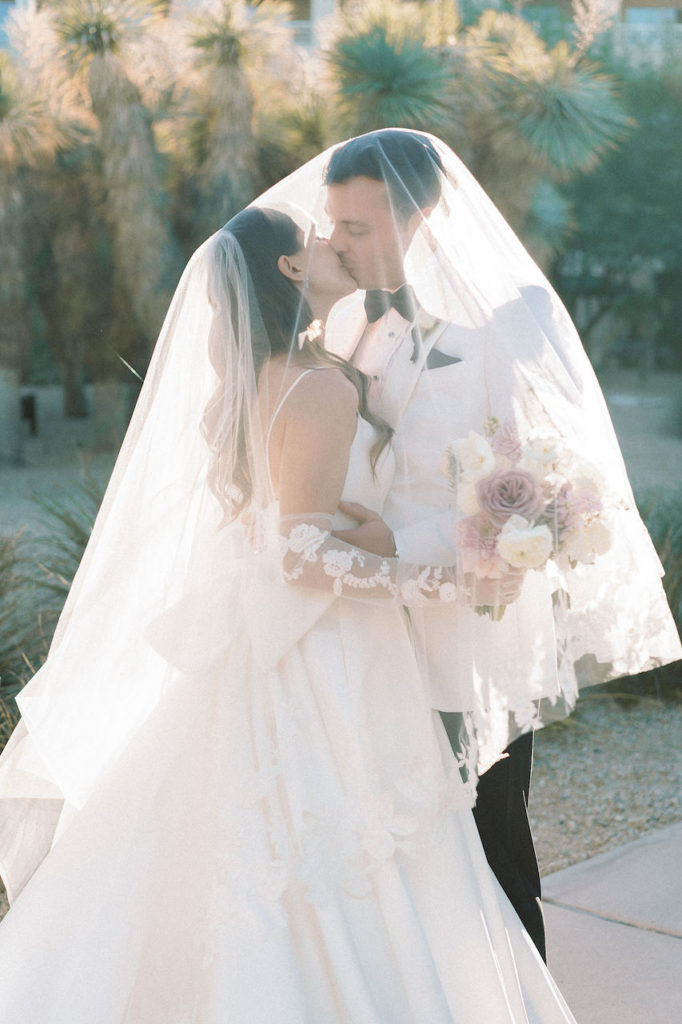 Groom and bride kissing under bride's veil outside with desert background.