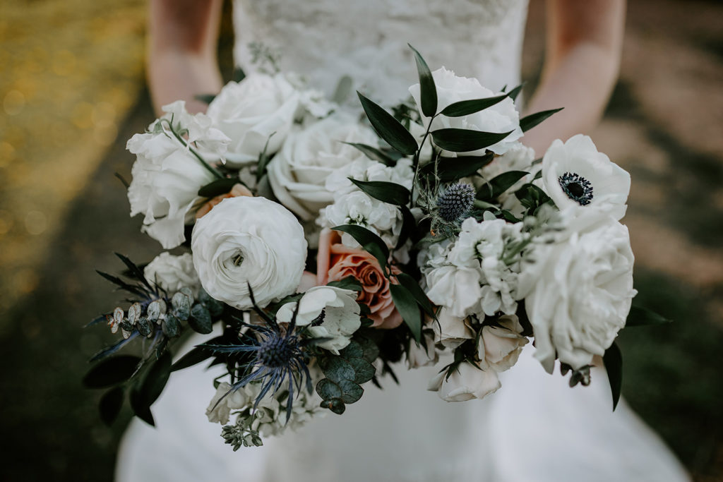Bouquet of white flowers including roses, anemone, and blue thistle with greenery.