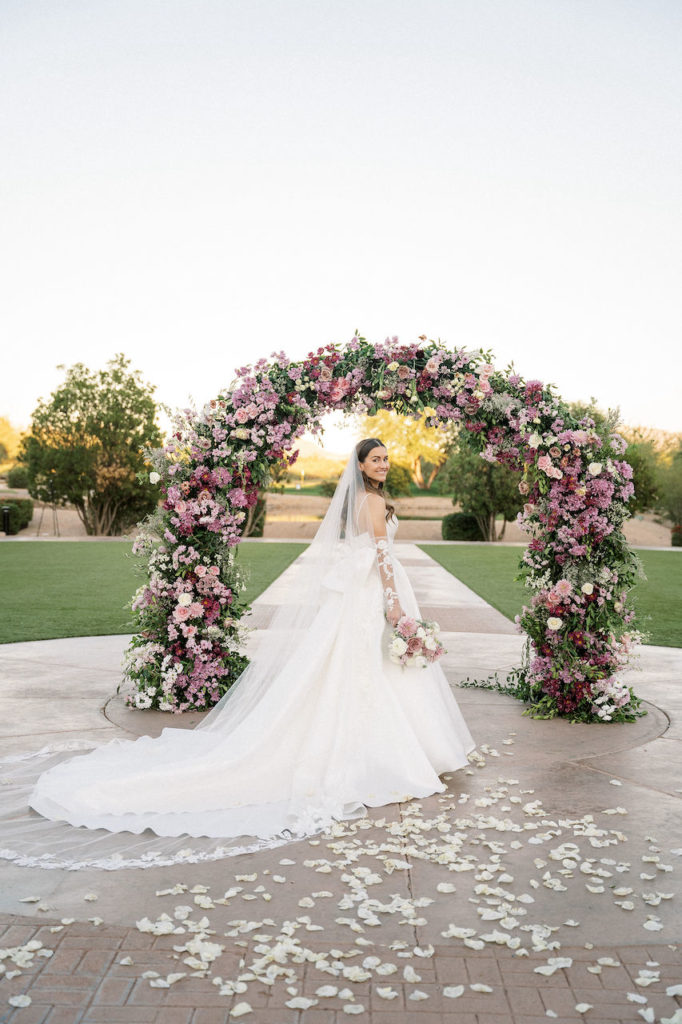 Bride looking over shoulder standing in front of wedding ceremony arch and holding bouquet.