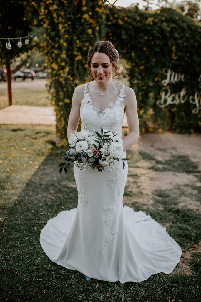 Bride standing outside, smiling, looking down at bouquet of white and pink flowers.