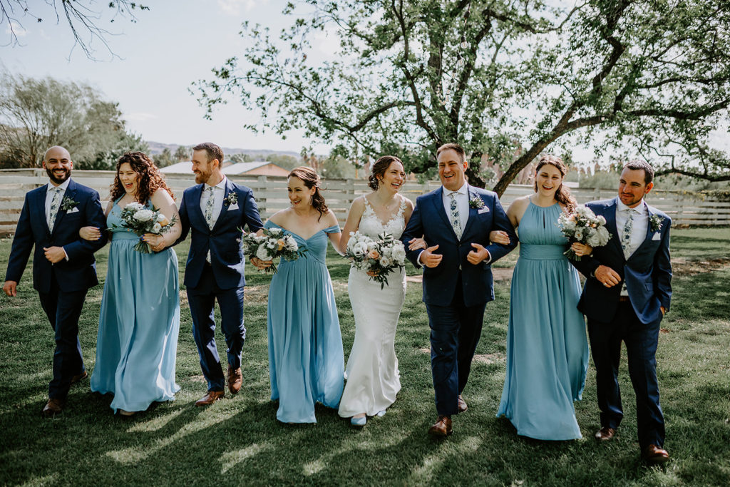 Bridal party walking with linked arms outdoors in grass field.