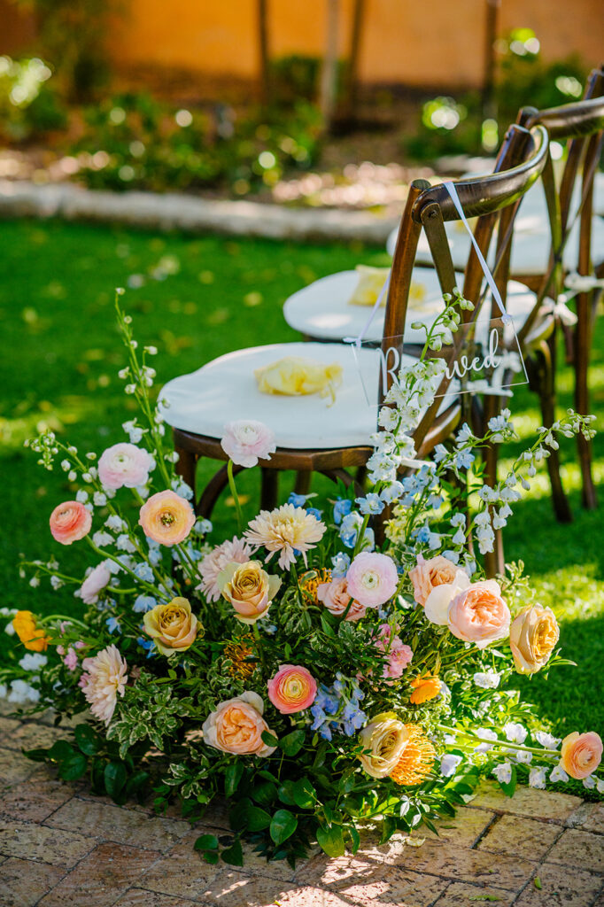 Floral ground arrangements in wedding ceremony aisle next to chairs of pink, peach, and yellow flowers.