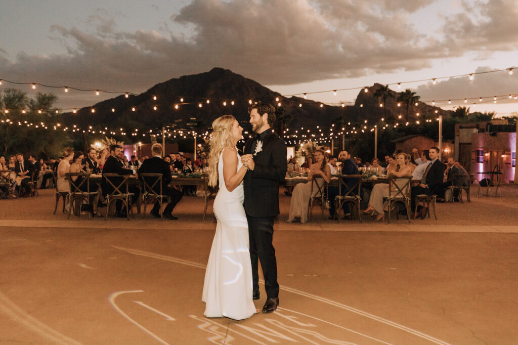 Bride and groom dancing outside at wedding reception in evening with hanging overhead lights and mountains in distance.