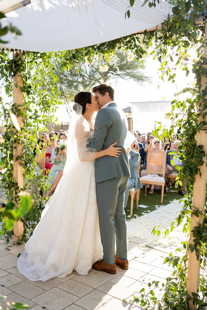 Bride and groom kissing during wedding ceremony under wedding arch decorated with greenery.