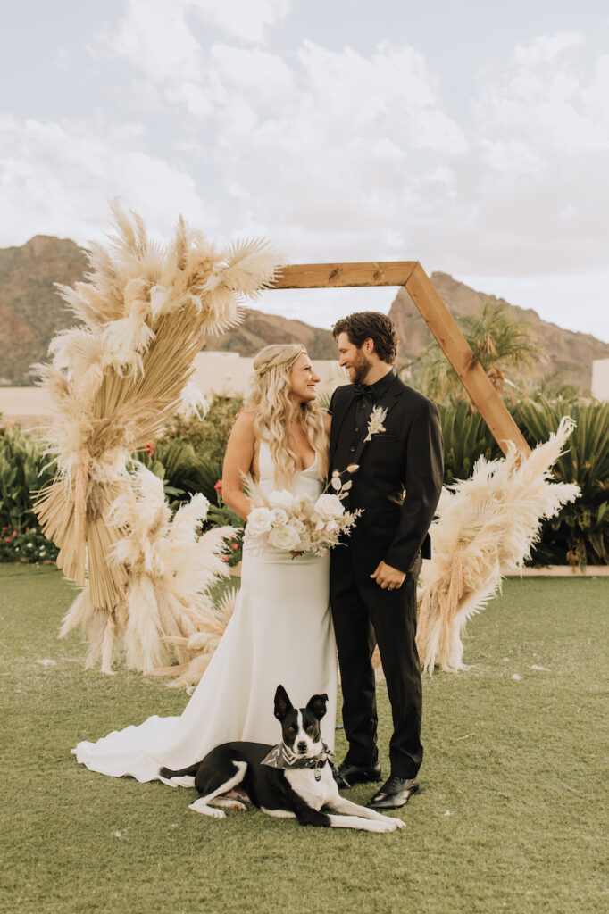 Bride and groom smiling at each other in front of hexagon arch with black and white dog laying down in front of them.