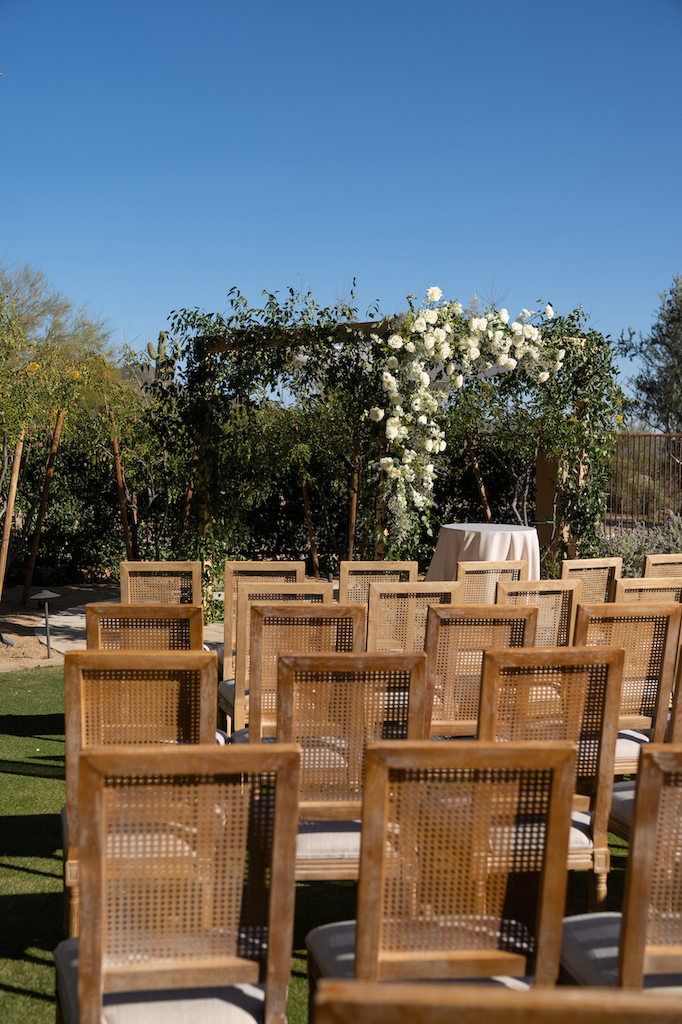 Outdoor wedding ceremony at DC Ranch with brown wooden guest chairs and square arch at front, decorated with greenery and white flowers.