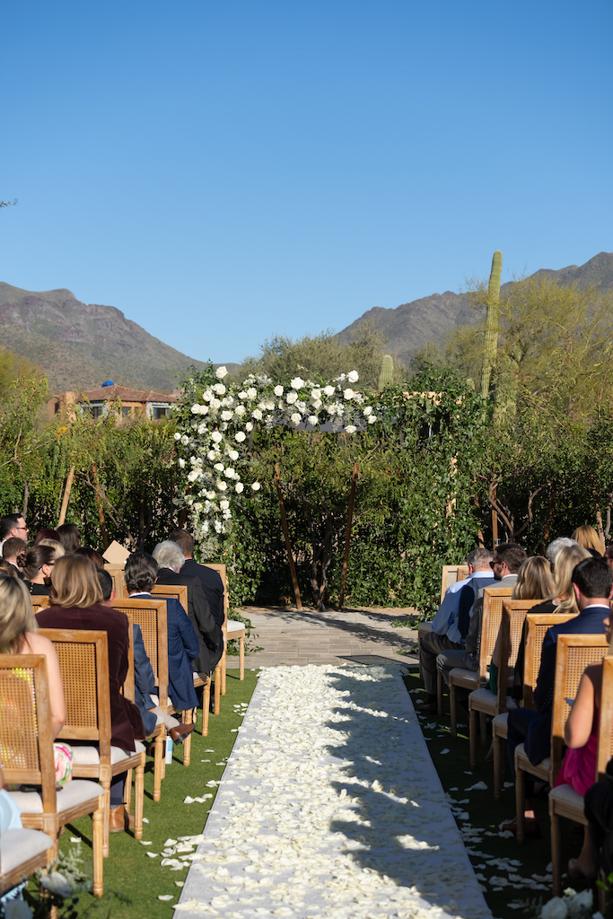 Wedding ceremony arch with white flowers and greenery and aisle lined with white rose petals at outdoor DC Ranch wedding with desert landscape in the background.