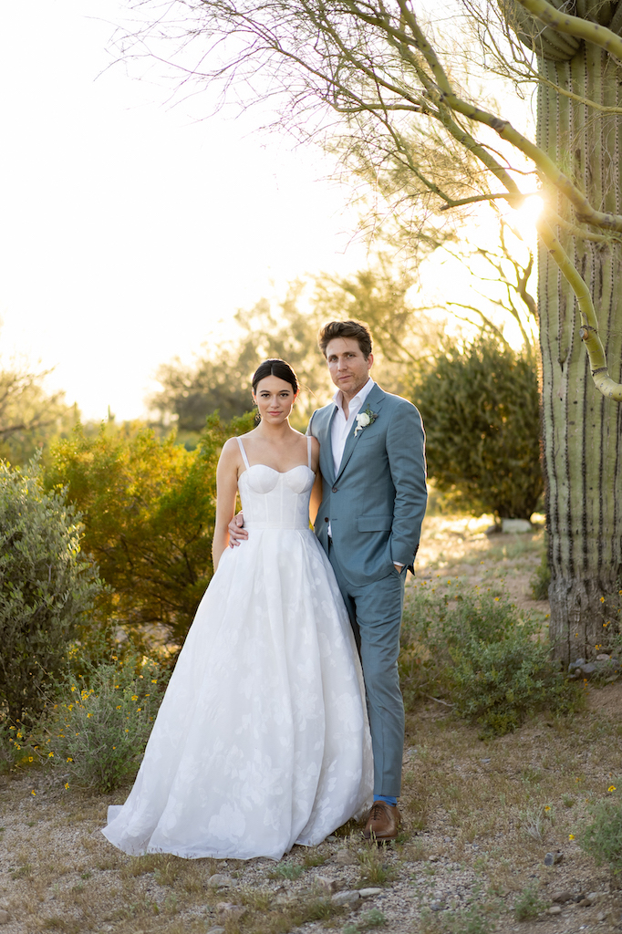 Bride and groom looking at camera, standing next to each other in desert landscape.