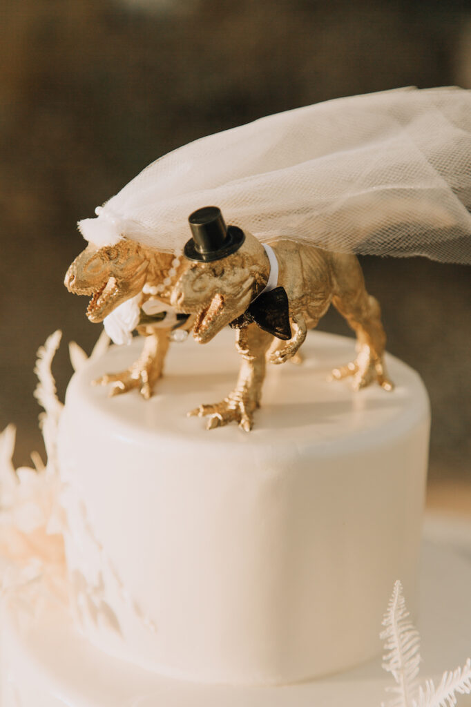 Gold dinosaur bride and groom cake toppers on white tiered wedding cake.