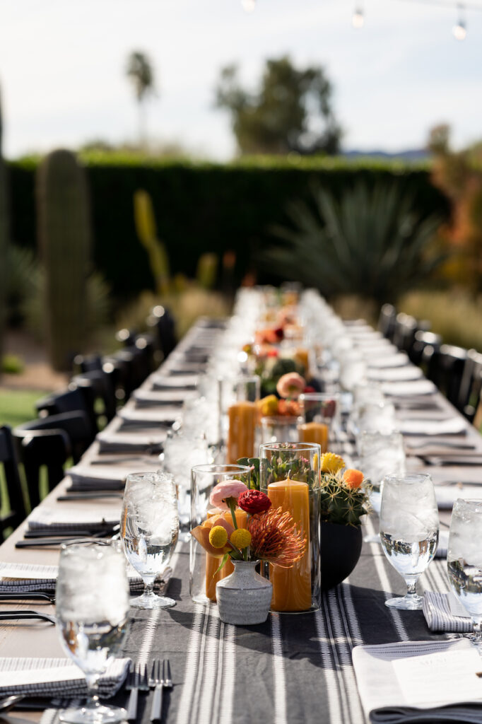 Long rectangular reception table with black table runner, place setting, amber pillar candles, potted succulents and bud vases.