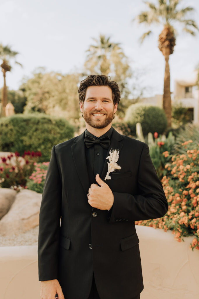 Groom smiling, holding onto black suit lapel he is wearing with dried floral boutonniere.