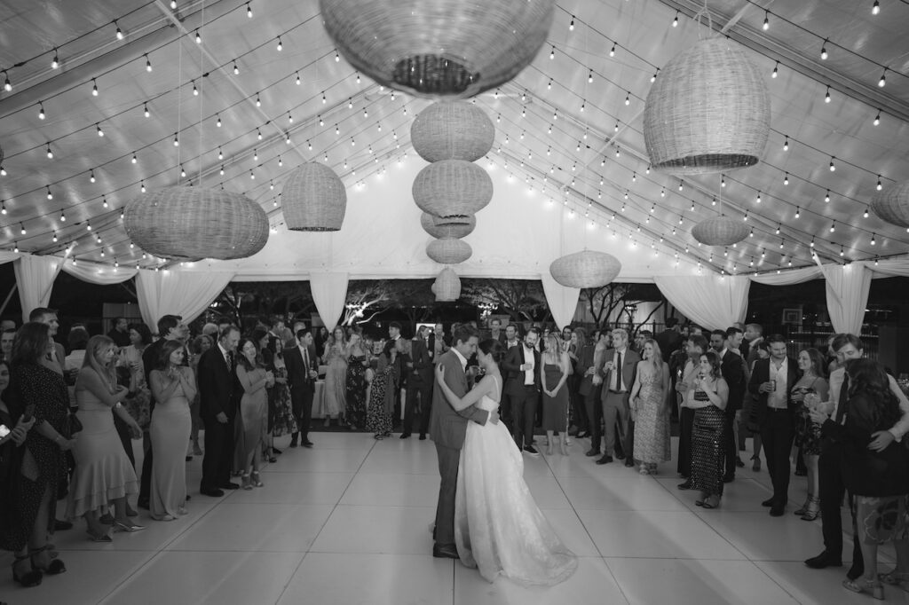 Wedding reception tent with woven handing chandeliers and bride and groom dancing in center with guests around them.