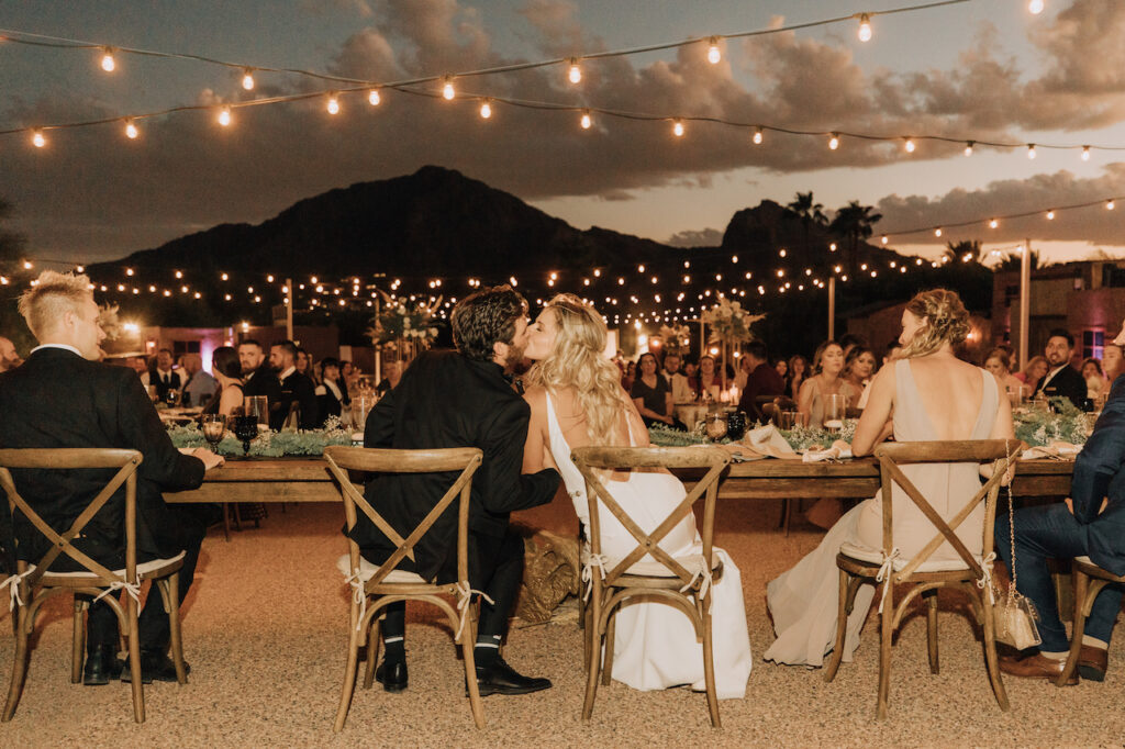 Bride and groom kissing while sitting at head table at outdoor wedding reception with mountains in distance.