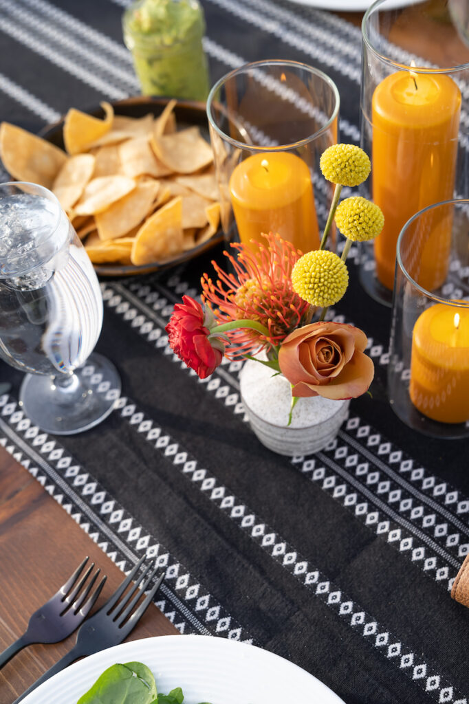 Bud vases of yellow, peach, and red flowers next to amber colored pillar candles on a black table runner.