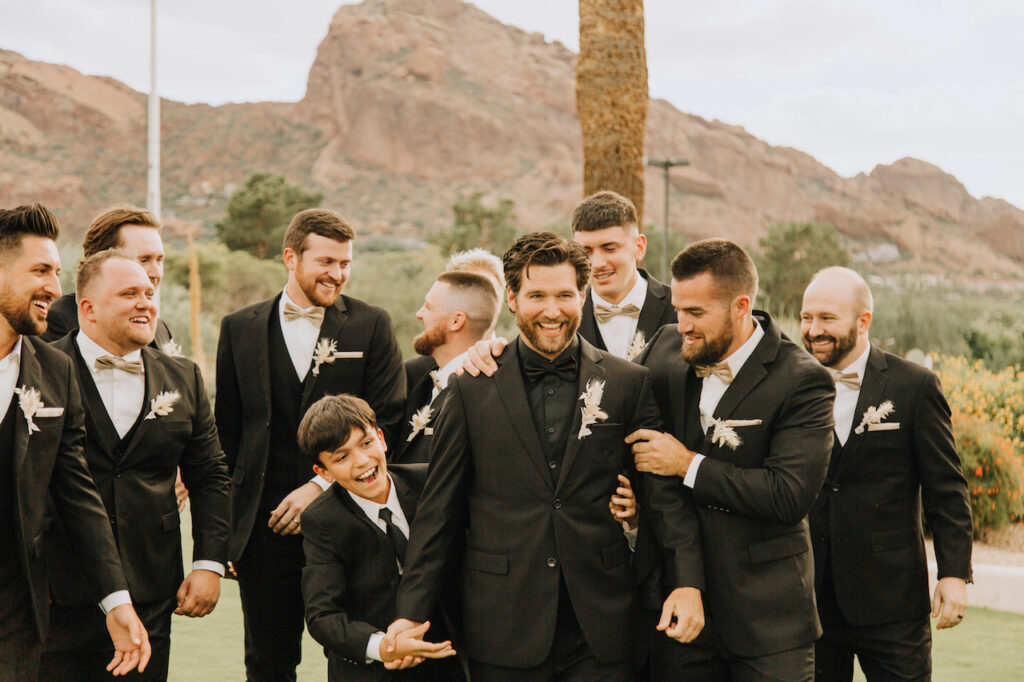 Groom with groomsmen and ring bearer in black suits outside in desert landscape setting.