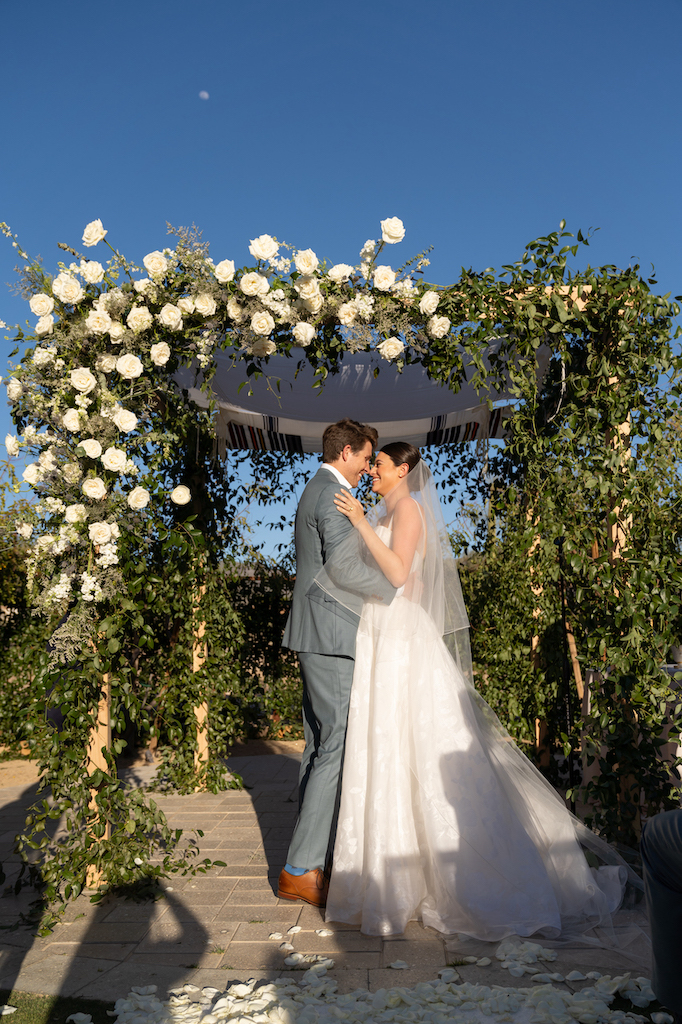 Bride and groom standing in front of ceremony wedding arch with white flowers and greenery attached to it.