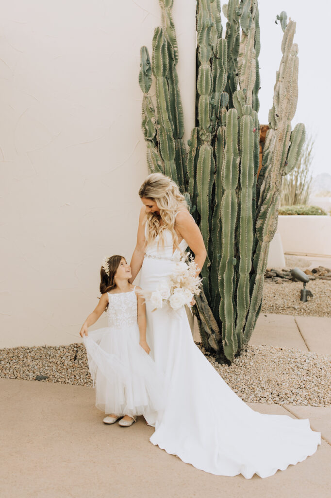 Bride standing with flower girl in white dress in front of large cactus, looking down and smiling at her.