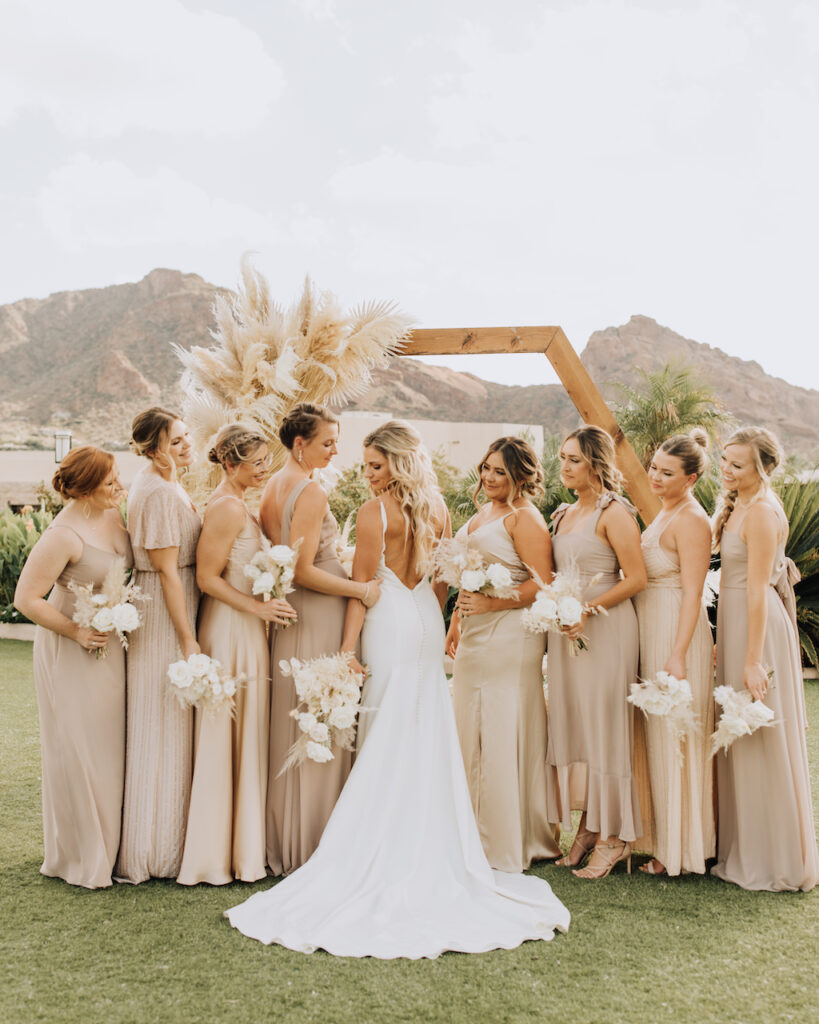 Bride with bridesmaids wearing taupe dresses standing in line outside in ceremony space.