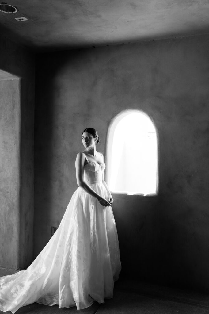 Bride wearing wedding dress, standing next to curved window, looking back over her shoulder.