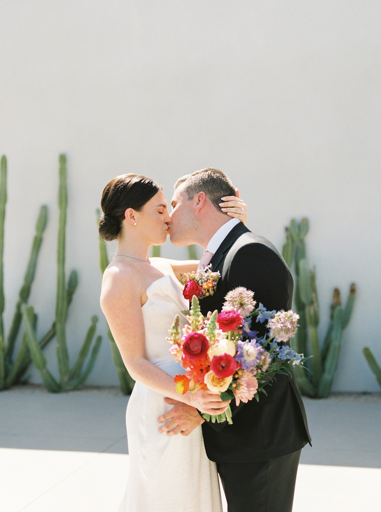 Bride and groom kissing in front of white wall with cacti in front of it.