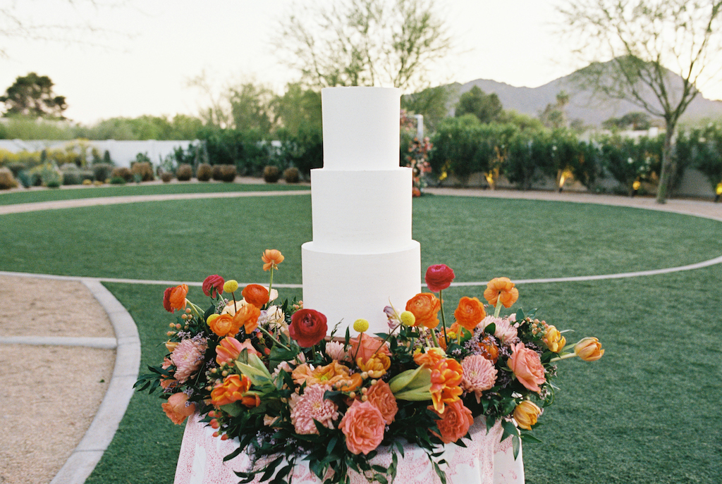 Three tiered white wedding cake with floral around it on table.