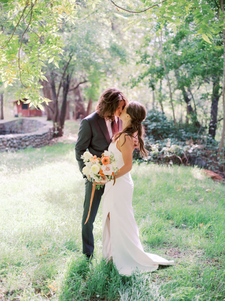 Bride and groom kissing in grass field with forest and short stone wall in background.