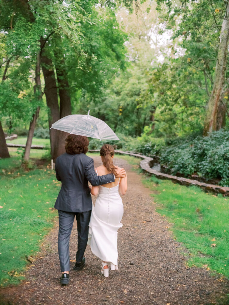 Bride and groom walking down gravel path under clear umbrella.