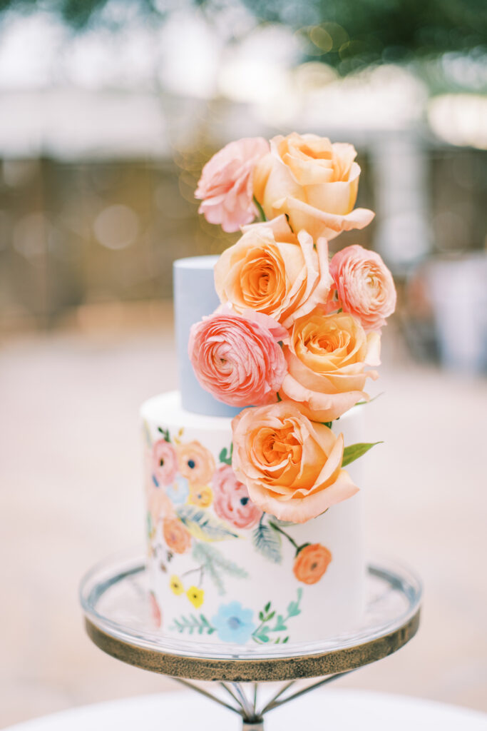 Two tiered wedding cake with blue top and floral painted white bottom with fresh flowers added.