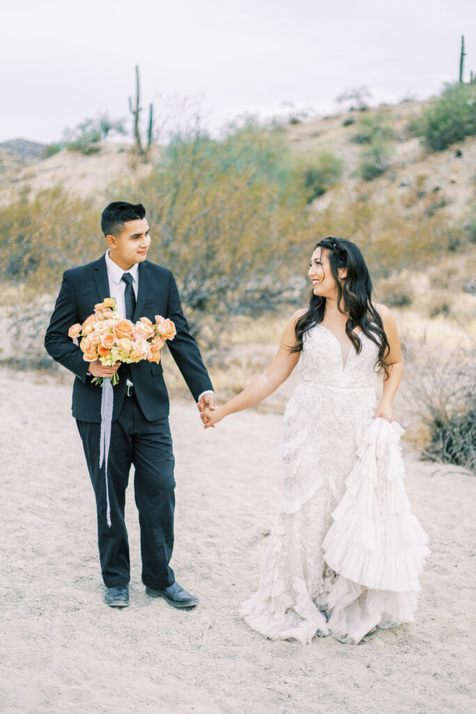 Bride and groom holding hands and walking in desert, groom holding her bouquet.