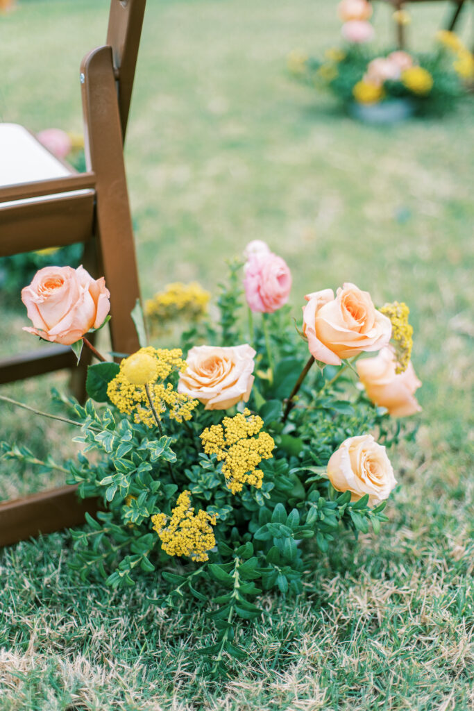 Ground floral arrangement at outdoor wedding ceremony of yellow yarrow and peach roses with greenery.
